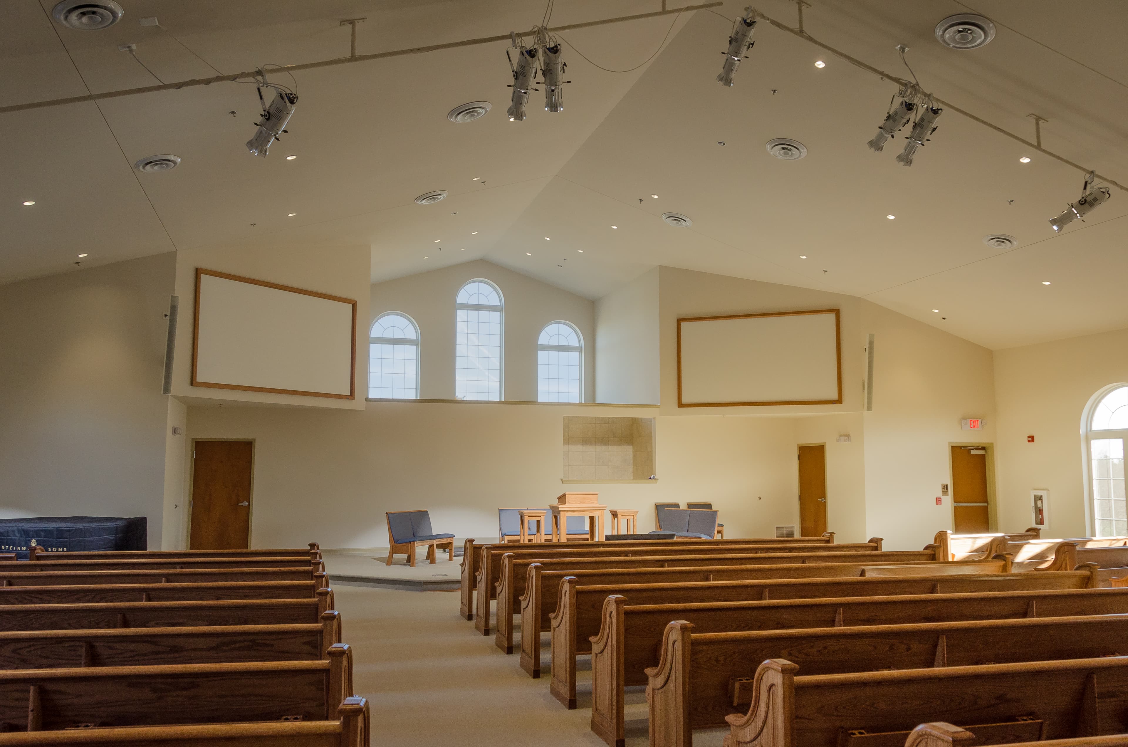 Church sanctuary interior with wooden pews, vaulted ceiling, arched windows, and mounted projection screens